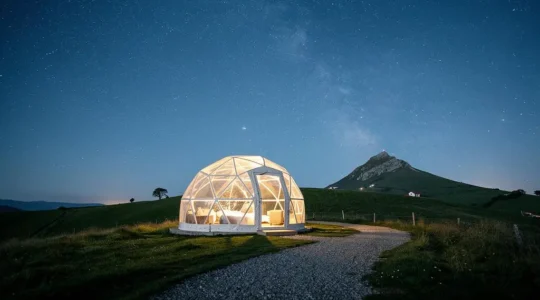 Bulle d'hébergement transparente installée sur une colline verdoyante du Pays Basque, sous un ciel étoilé avec le sommet de la Rhune en silhouette au loin